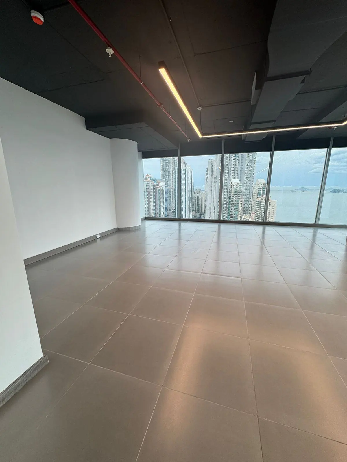 Empty office space with gray tiled floor, white walls, and black ceiling. Large windows offer a city view.