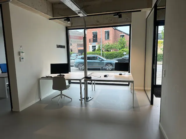 Bright office space with white walls, gray floors, and large windows. Desks with computers and chairs face the street view. "Lounge" sign on the wall.