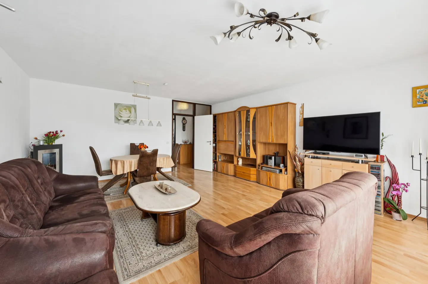 Living room with brown sofas, wood floors, and white walls. A dining table and TV stand are also visible.