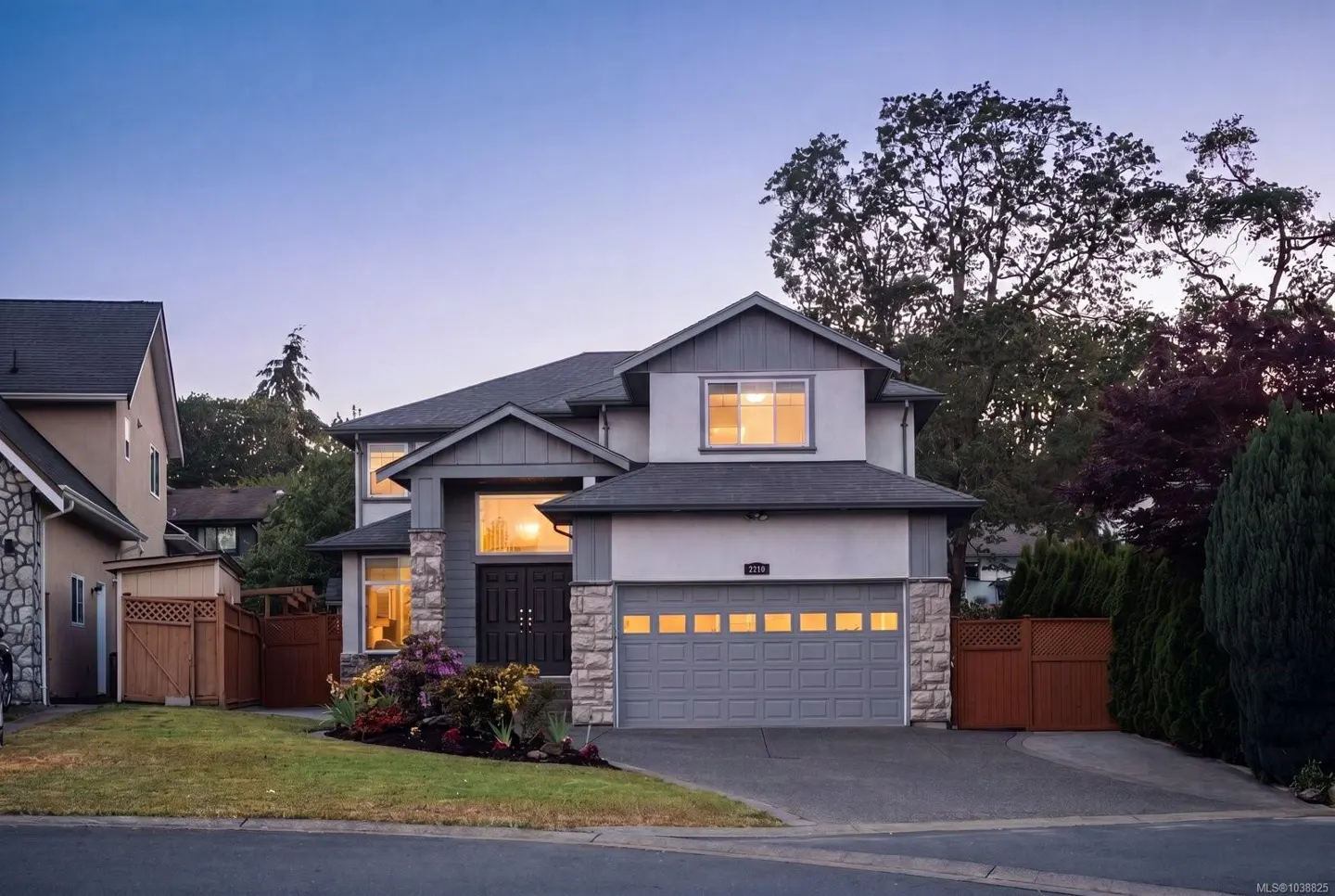 Two-story suburban home at dusk with a grey garage, stone accents, and warm interior lighting glowing through windows.
