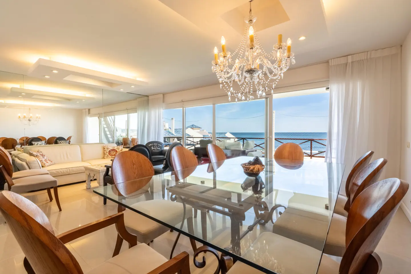 Bright, airy dining room with glass table, wood chairs, and ocean view. Chandelier hangs above. White sofa and chairs in background.