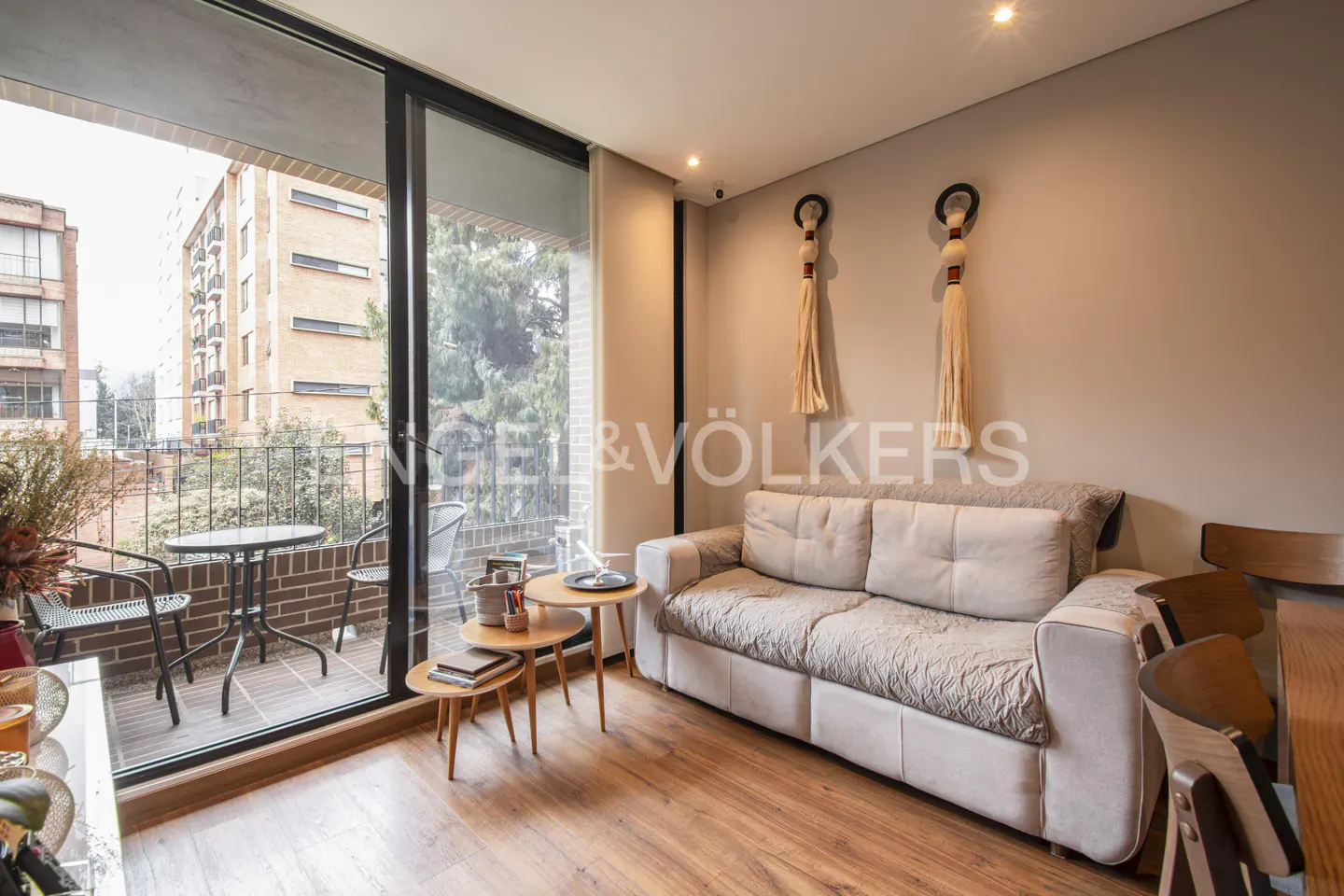 Living room with a beige sofa, wooden floor, and a balcony with a table and chairs. Two decorative tassels hang on the wall.