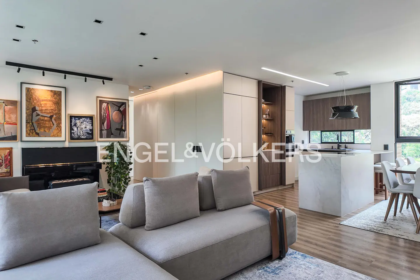 Open-concept living space with a gray sofa, piano, art, and kitchen island. The room has light wood floors and natural light.