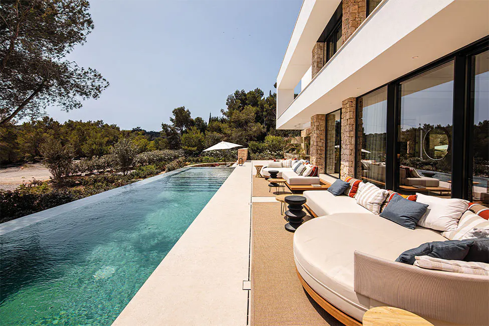 Modern villa with an infinity pool and outdoor lounge area. The building has stone accents and large windows. The pool overlooks lush greenery.