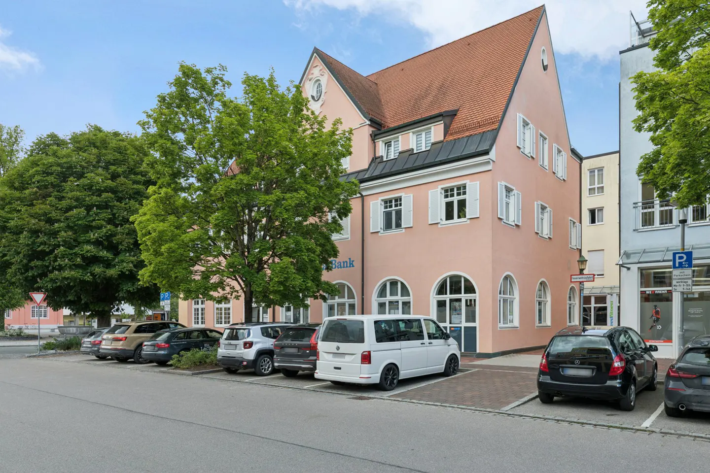A three-story pink building with a red roof and white shutters, featuring a parking lot with several cars in front.