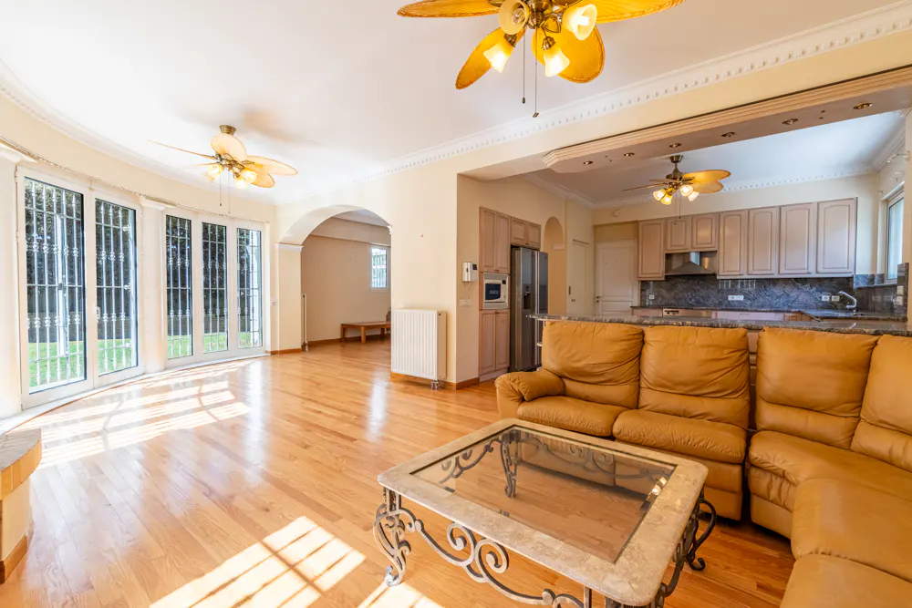 Open-concept living room with hardwood floors, tan sofa, glass coffee table, and kitchen in the background. Ceiling fans overhead.