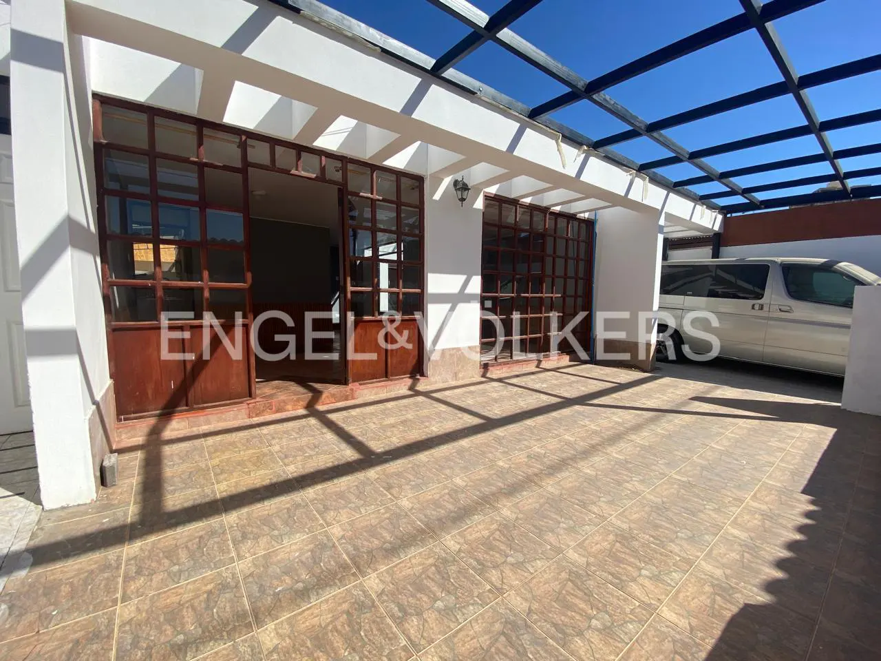 Exterior view of a white building with brown framed windows and a black pergola. A silver van is parked in the background. The ground is tiled.