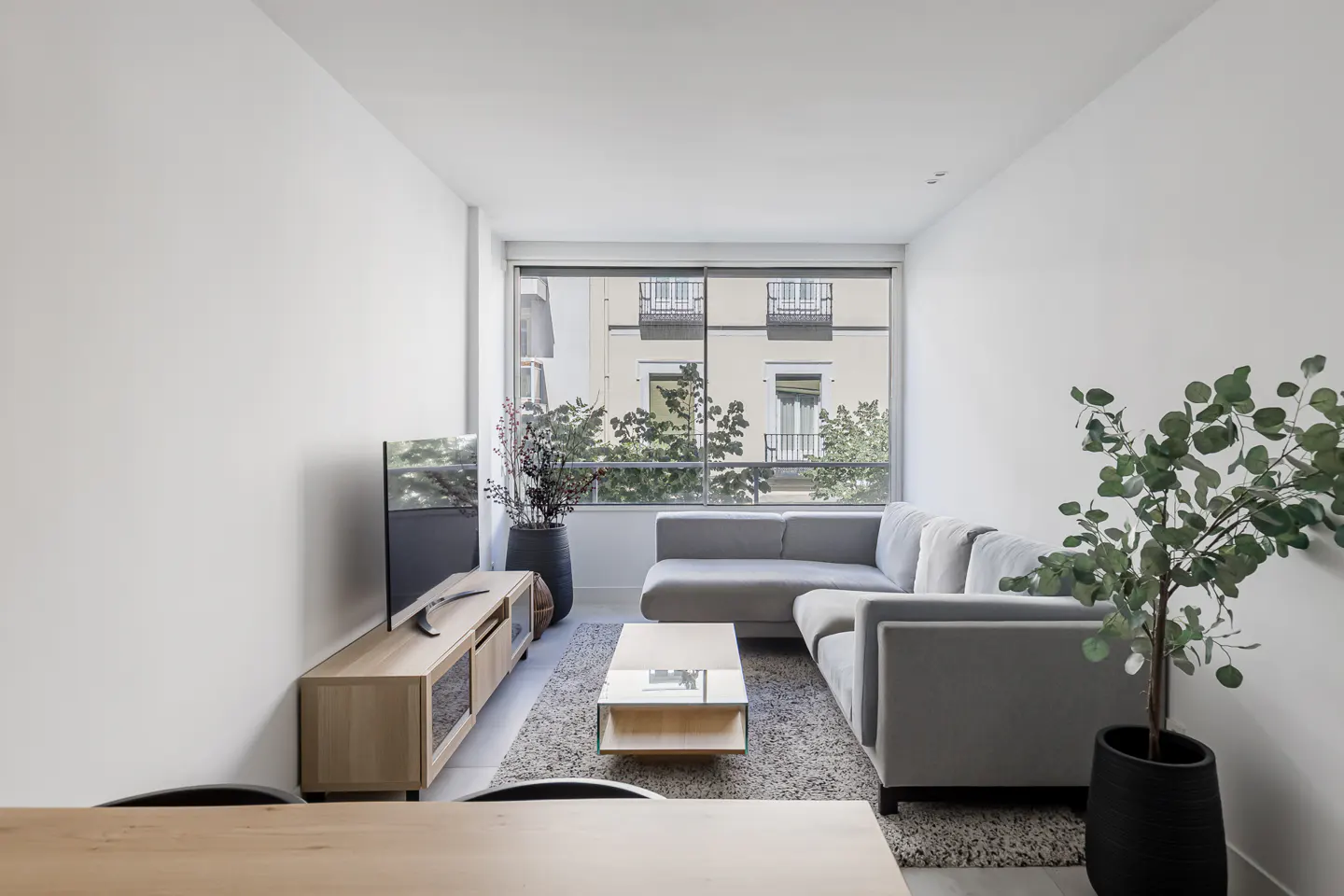 Modern living room with a grey sectional sofa, light wood media console, glass coffee table, and potted plants.