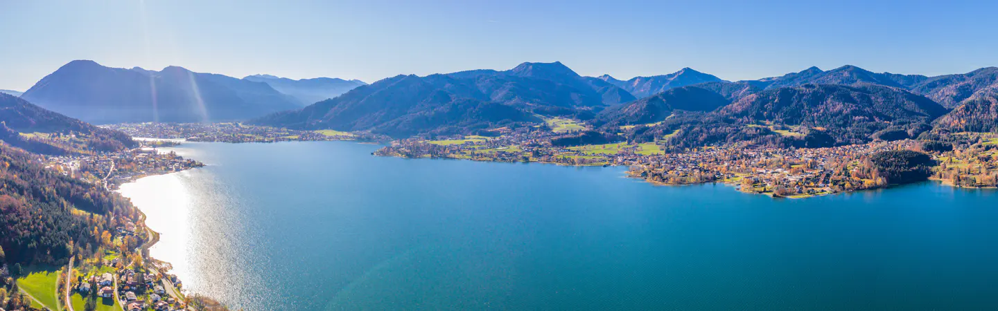 Aerial view of a blue lake surrounded by green mountains and small towns under a clear blue sky.