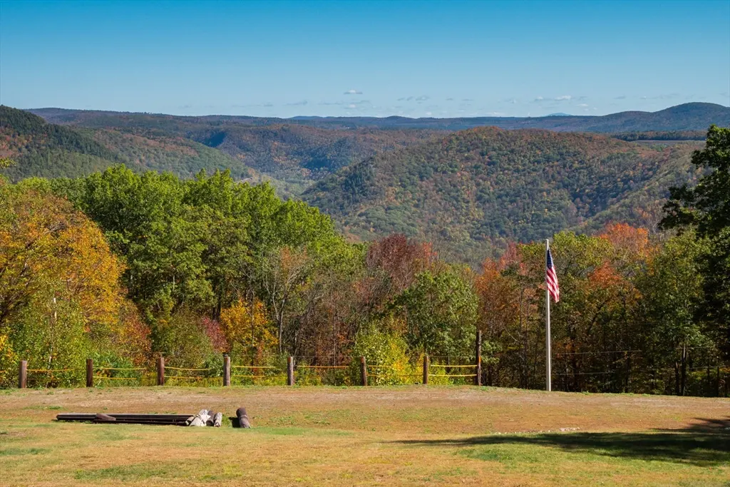 Scenic view of rolling hills with fall foliage under a blue sky. An American flag stands near a wooden fence on a grassy lawn.