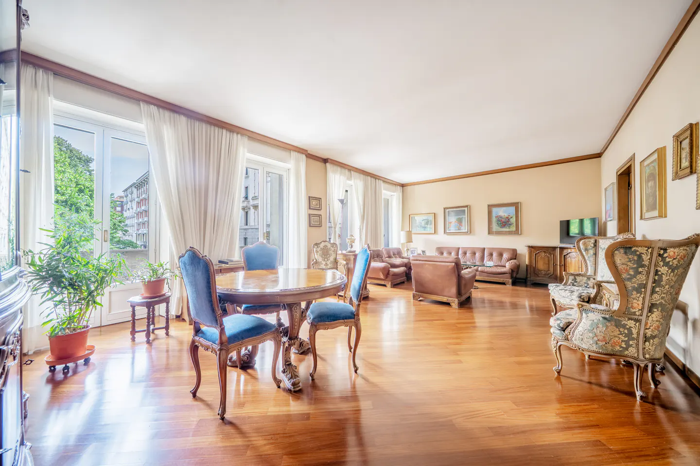 Spacious living and dining room with hardwood floors, blue velvet chairs, leather sofas, and large windows with white drapes.