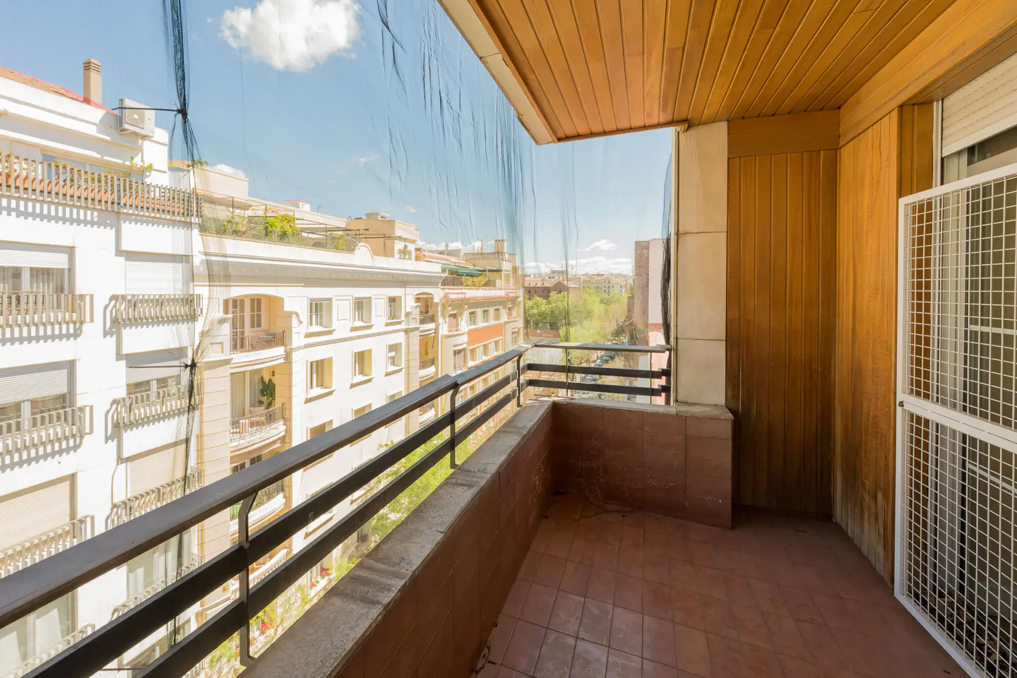 Balcony with brown tile floor, black metal railing, and a view of city buildings through a protective net.