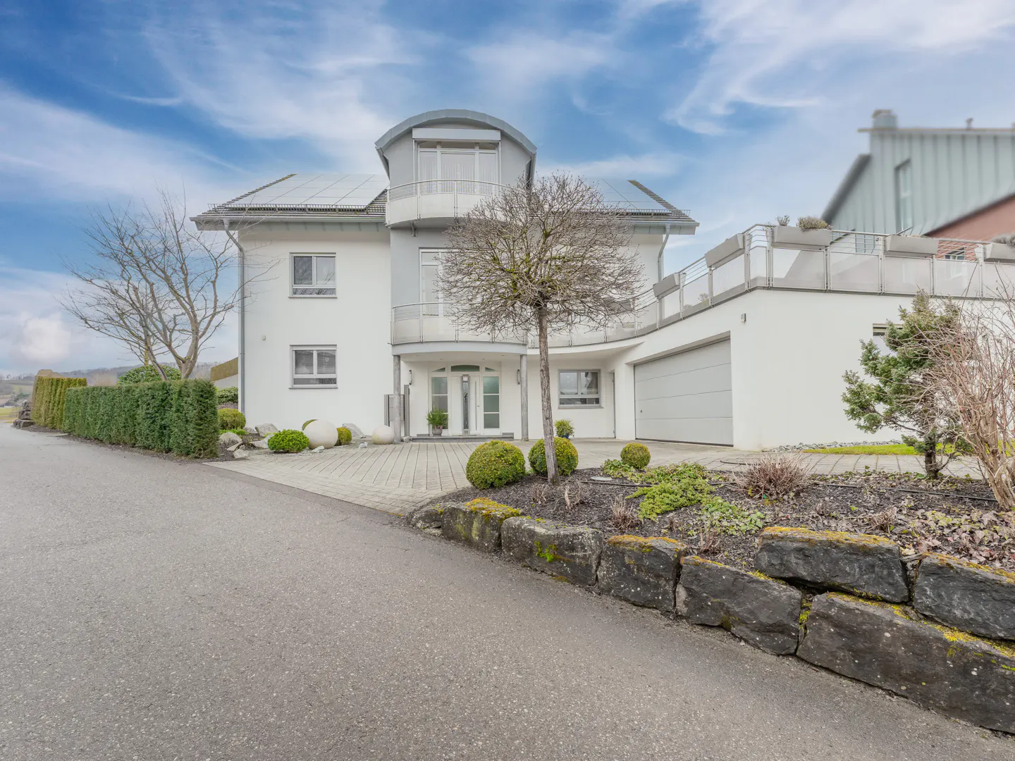 Two-story white house with a curved balcony, a garage, and a stone retaining wall in the front yard.