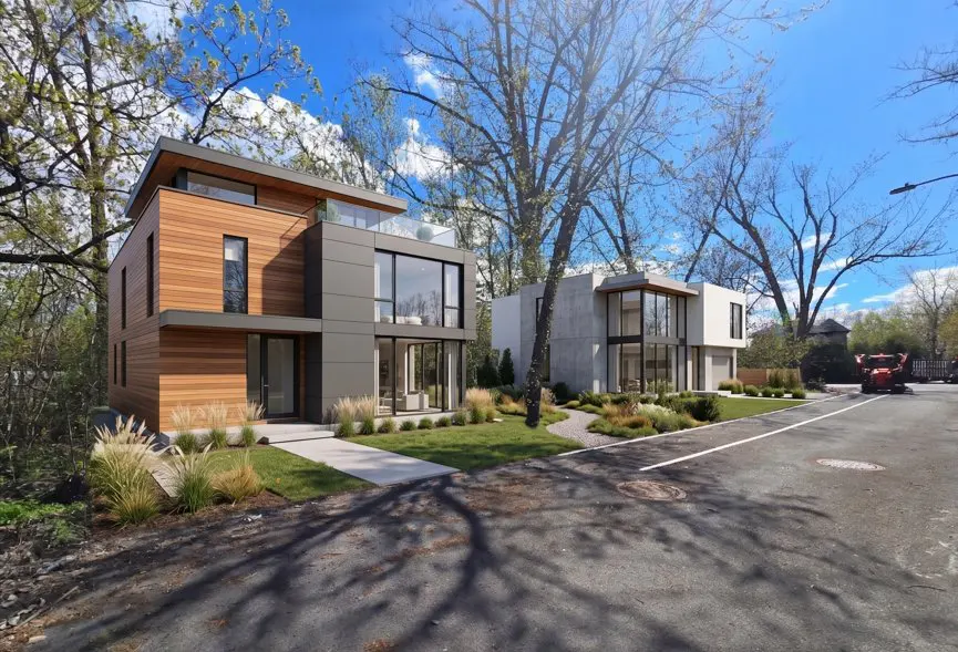 Two modern homes with large windows and wood and concrete exteriors sit on a tree-lined street under a blue sky.