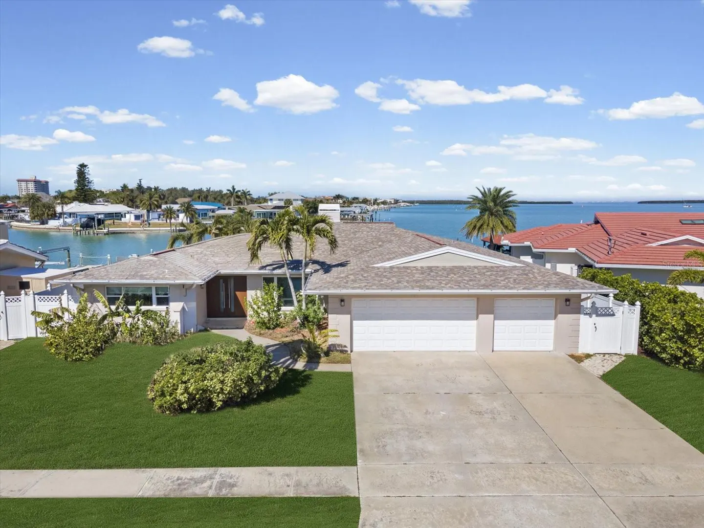 Waterfront home with a two-car garage, green lawn, and palm trees under a blue sky with fluffy white clouds.