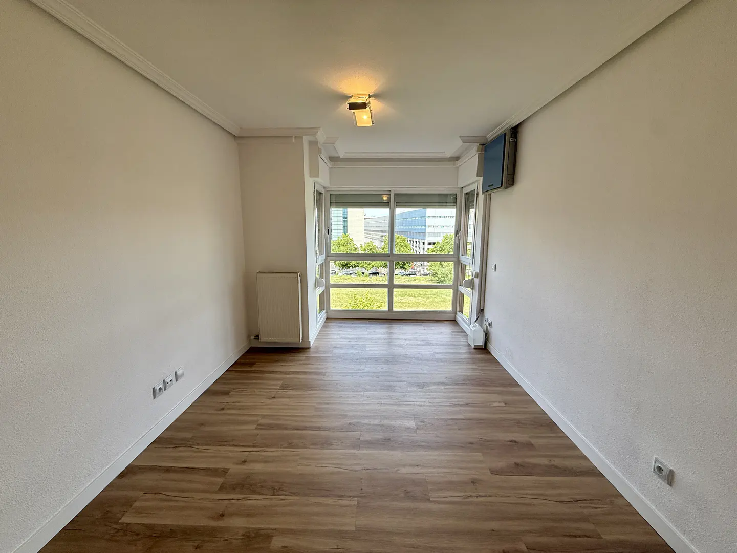 Empty room with wood floor, white walls, and a large window showing a green lawn and buildings.