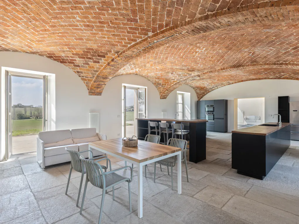 Open-plan living space with brick vaulted ceiling, stone floors, white walls, and a modern kitchen with black cabinets. A table and sofa are visible.
