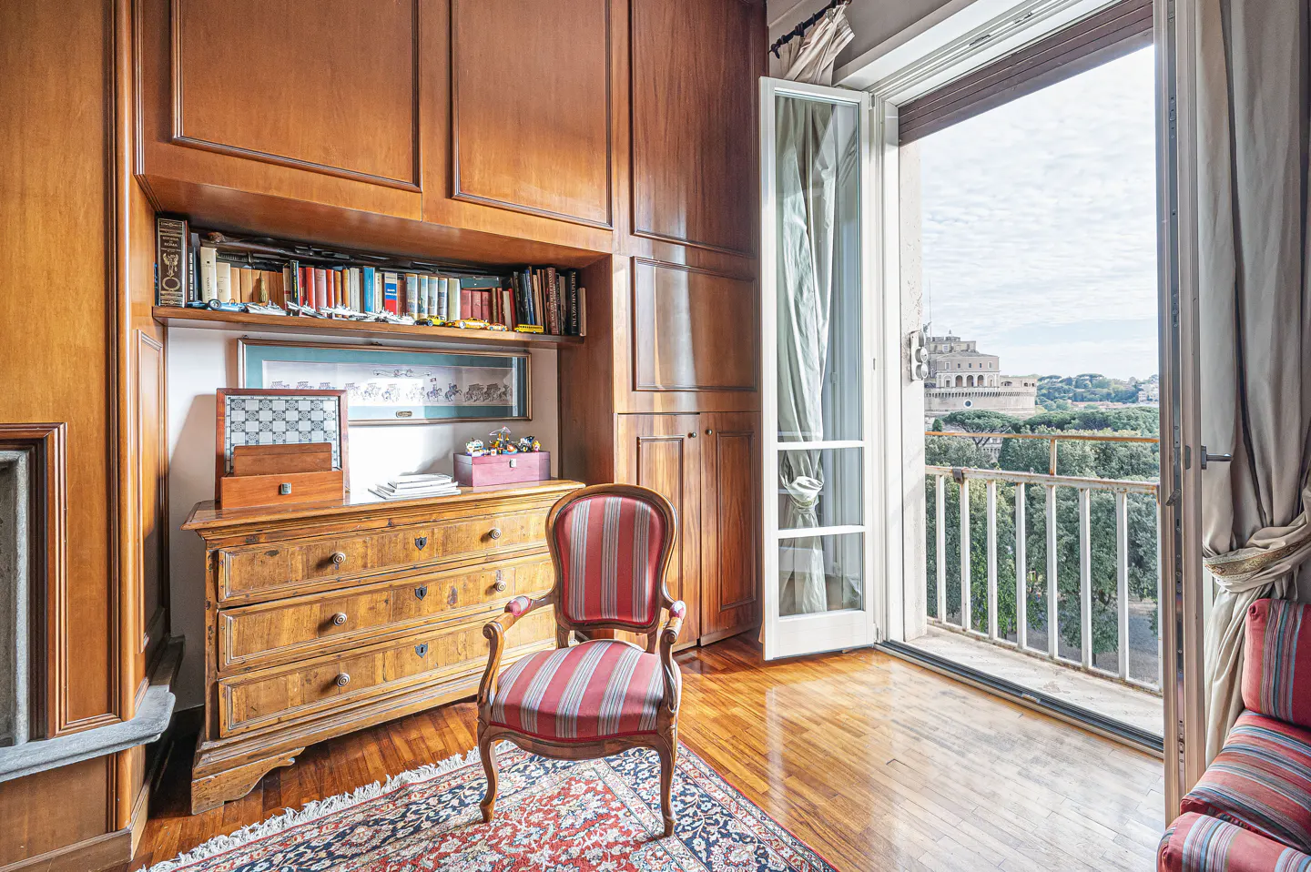 A room with wood paneling, a dresser with books, and a red and white striped chair. An open balcony door shows a view of a building.