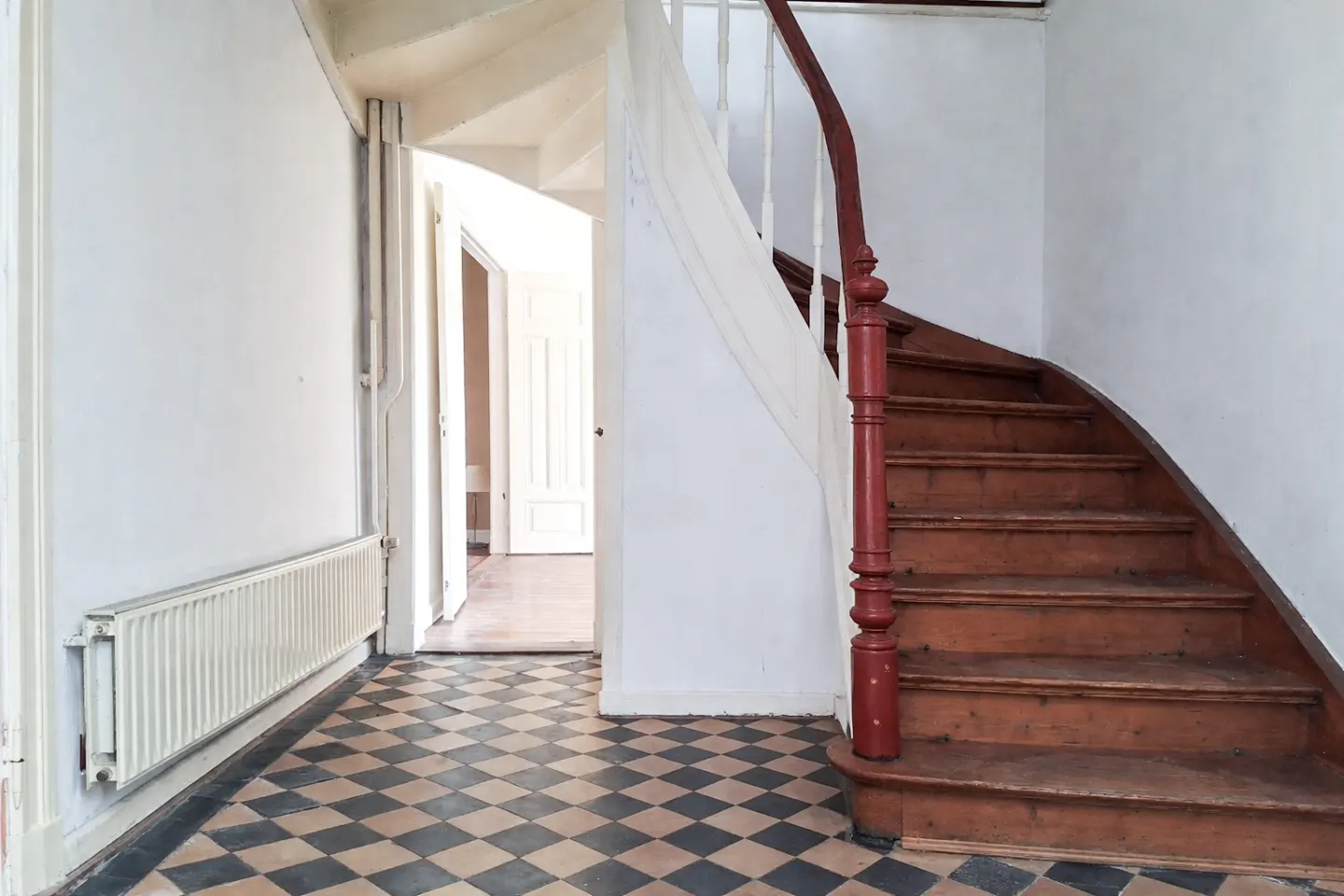 Interior view of a home's entryway with a wooden staircase, red banister, and black and beige checkered tile floor.