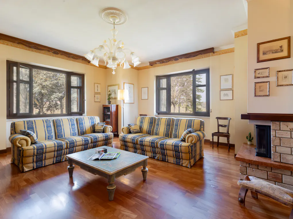 Living room with two blue and yellow striped sofas, a coffee table, and a fireplace. Two windows overlook trees.