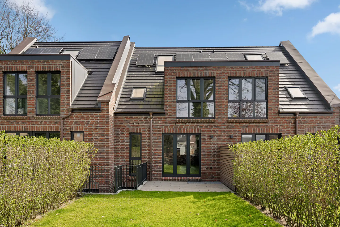 Rear view of a red brick house with black framed windows, a tiled roof with solar panels, and a green lawn.