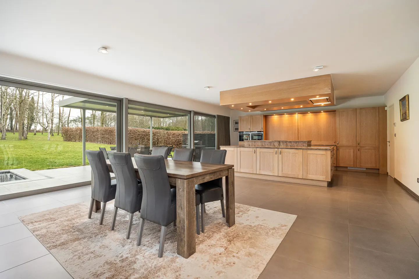 Open-concept kitchen and dining area with a wooden table, gray chairs, and a large window overlooking a green lawn.