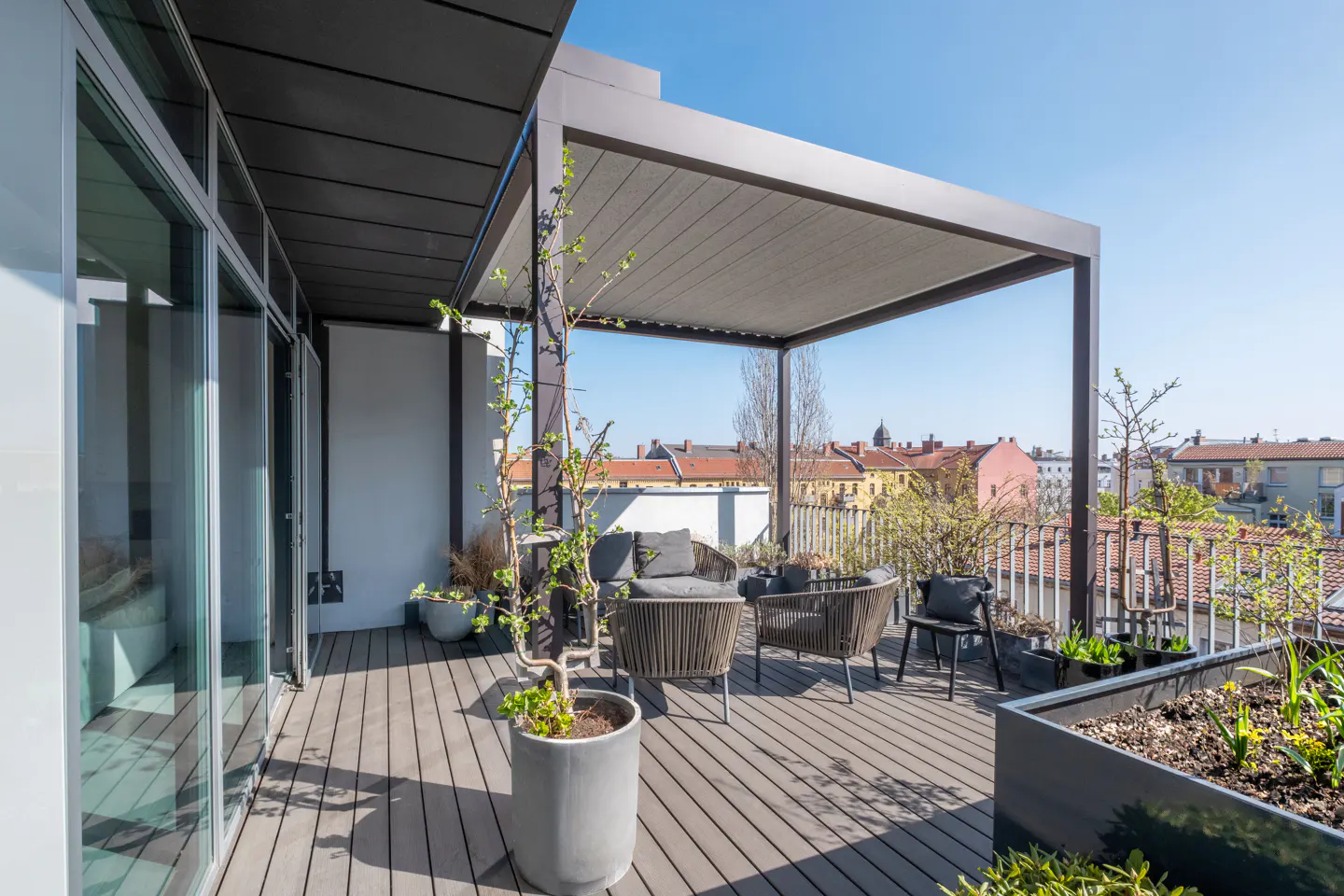Modern rooftop terrace with grey decking, outdoor lounge furniture, a pergola, and potted plants overlooking a city skyline.