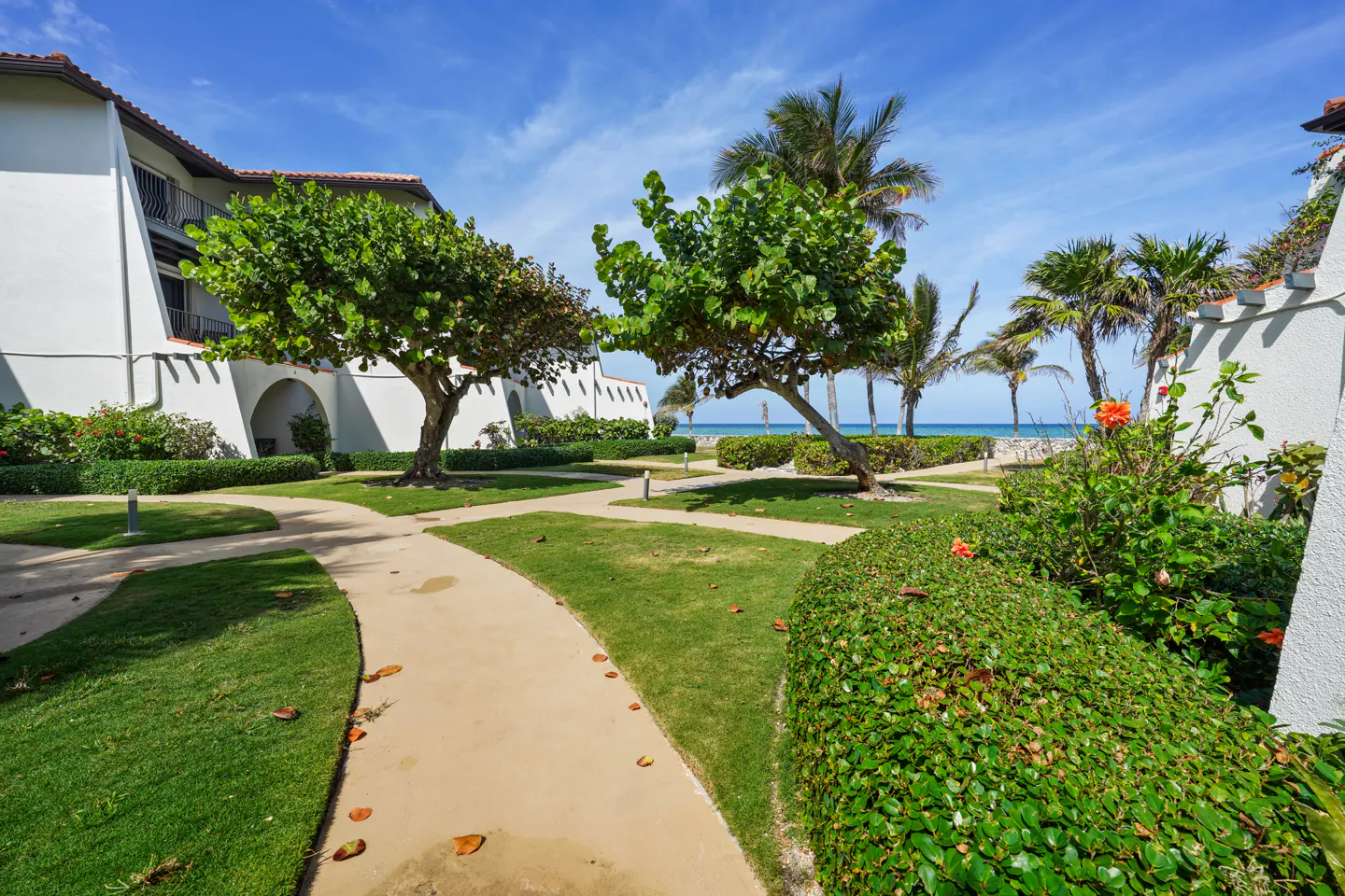 A winding path through a lush green lawn leads to a sandy beach and ocean view, framed by palm trees and white buildings.