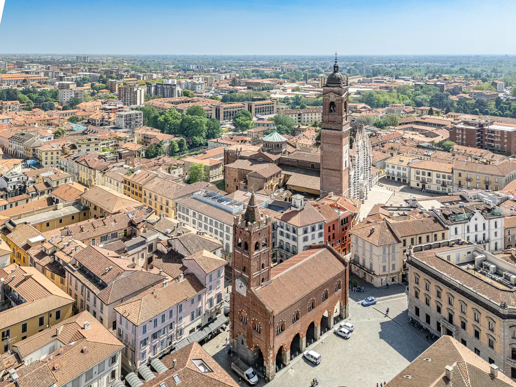 Aerial view of Monza, Italy, featuring the Duomo, bell tower, and surrounding buildings with red tile roofs under a blue sky.