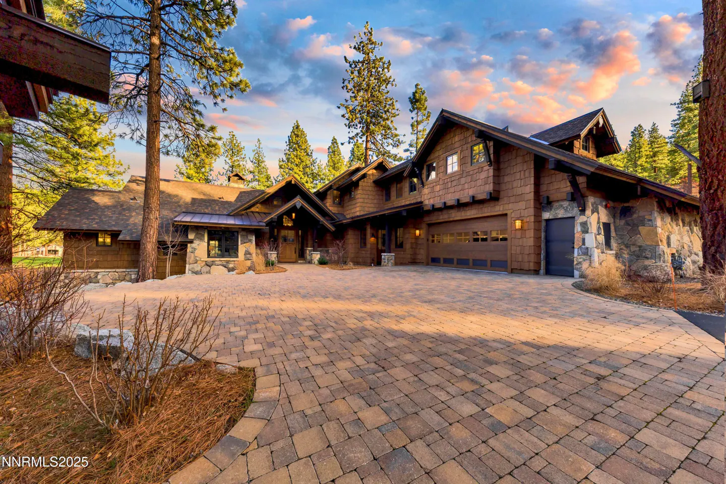 Exterior view of a large, brown, wood-shingled house with a stone facade and a brick driveway, surrounded by tall pine trees.