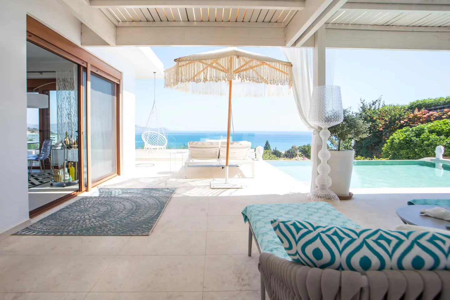 Luxury patio with ocean view. White pergola, lounge chairs, and a fringed umbrella. A pool and lush greenery are visible in the background.