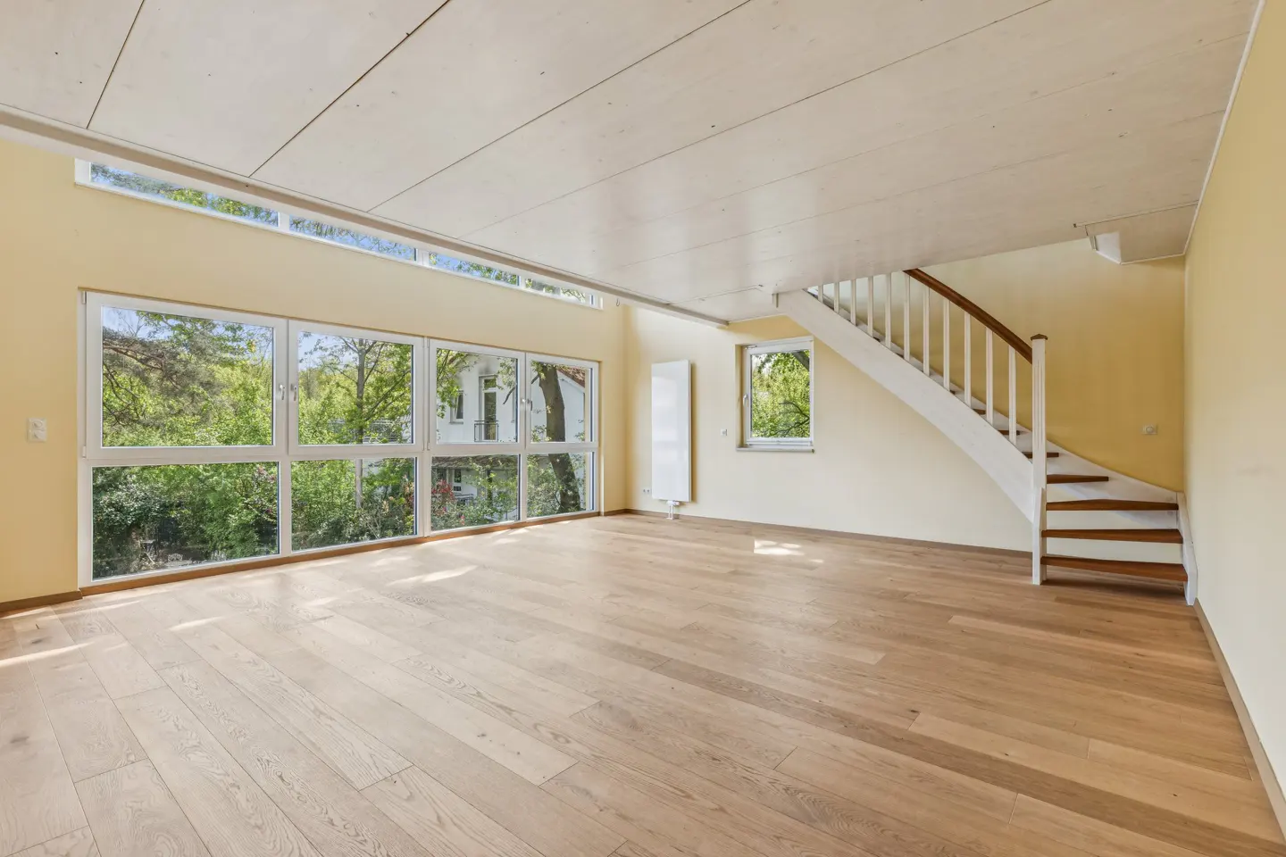 Bright, empty room with wood floors, yellow walls, and white ceiling. Large windows overlook trees. A white staircase with wood treads is on the right.