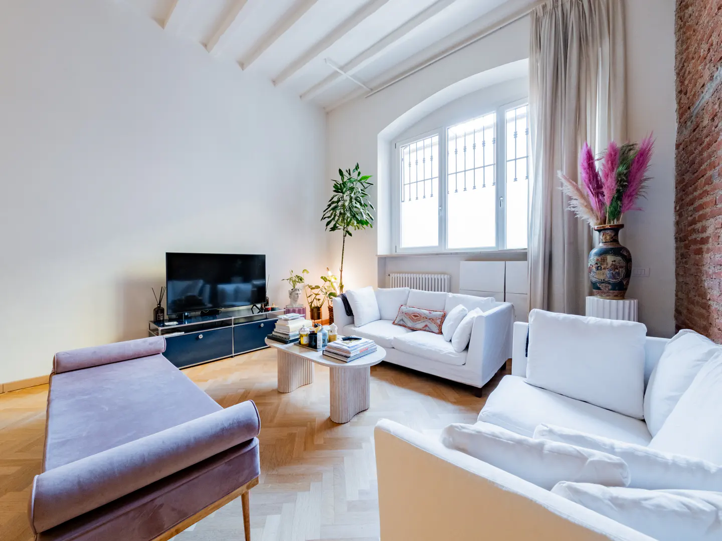Bright living room with white sofas, a lilac chaise lounge, and a TV on a blue cabinet. A large window and herringbone wood floors add to the room's appeal.