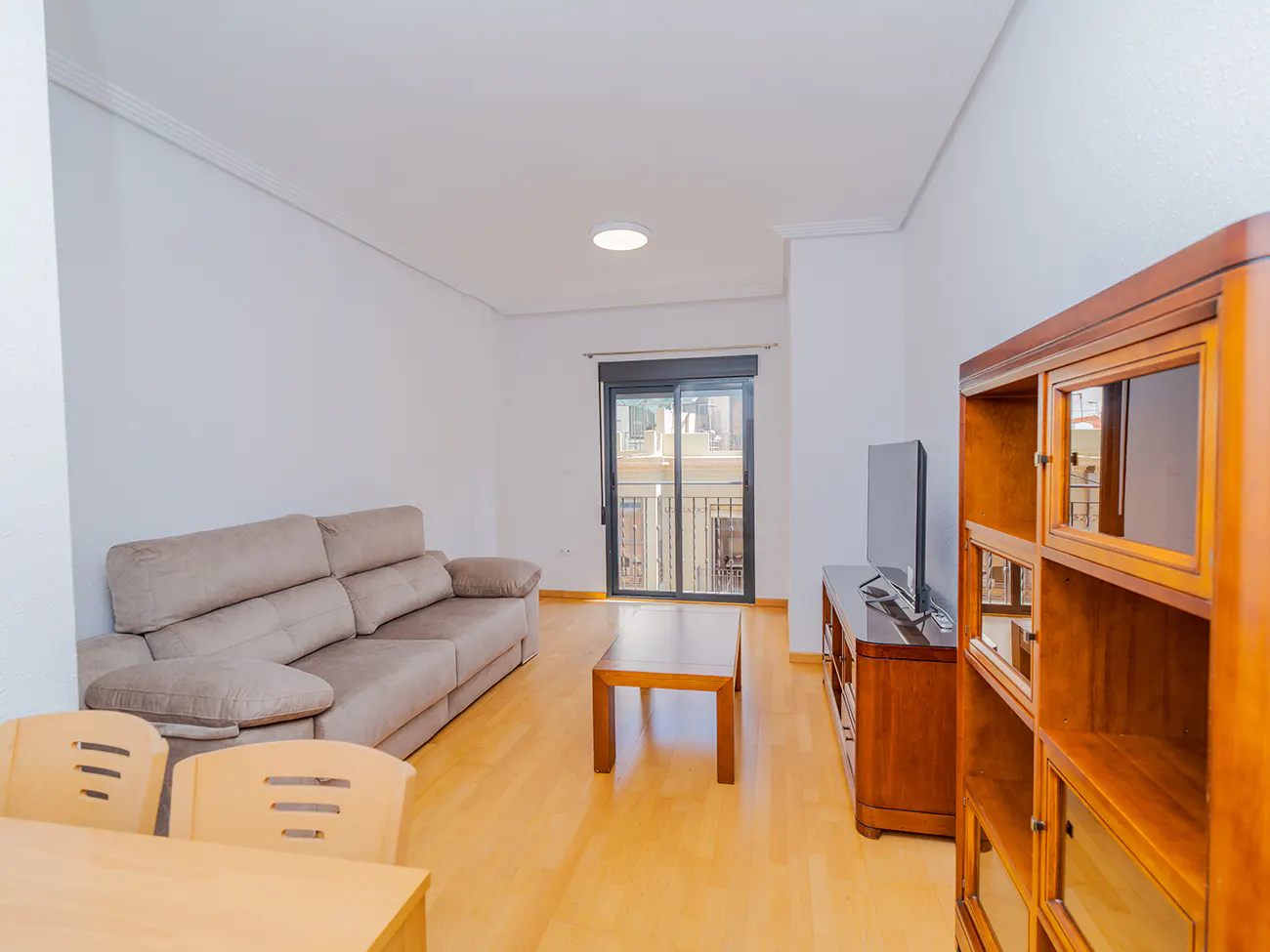 Bright living room with a beige sofa, wooden coffee table, TV, and a dark wood bookcase. A sliding glass door leads to a balcony.
