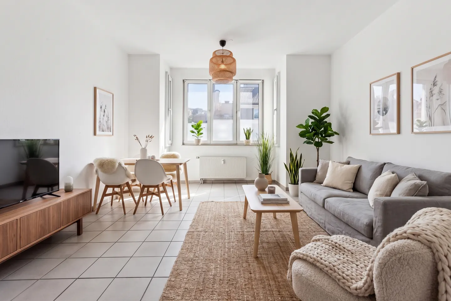 Bright living room with a gray sofa, wooden table, and woven rug. Plants and artwork add warmth to the white walls.