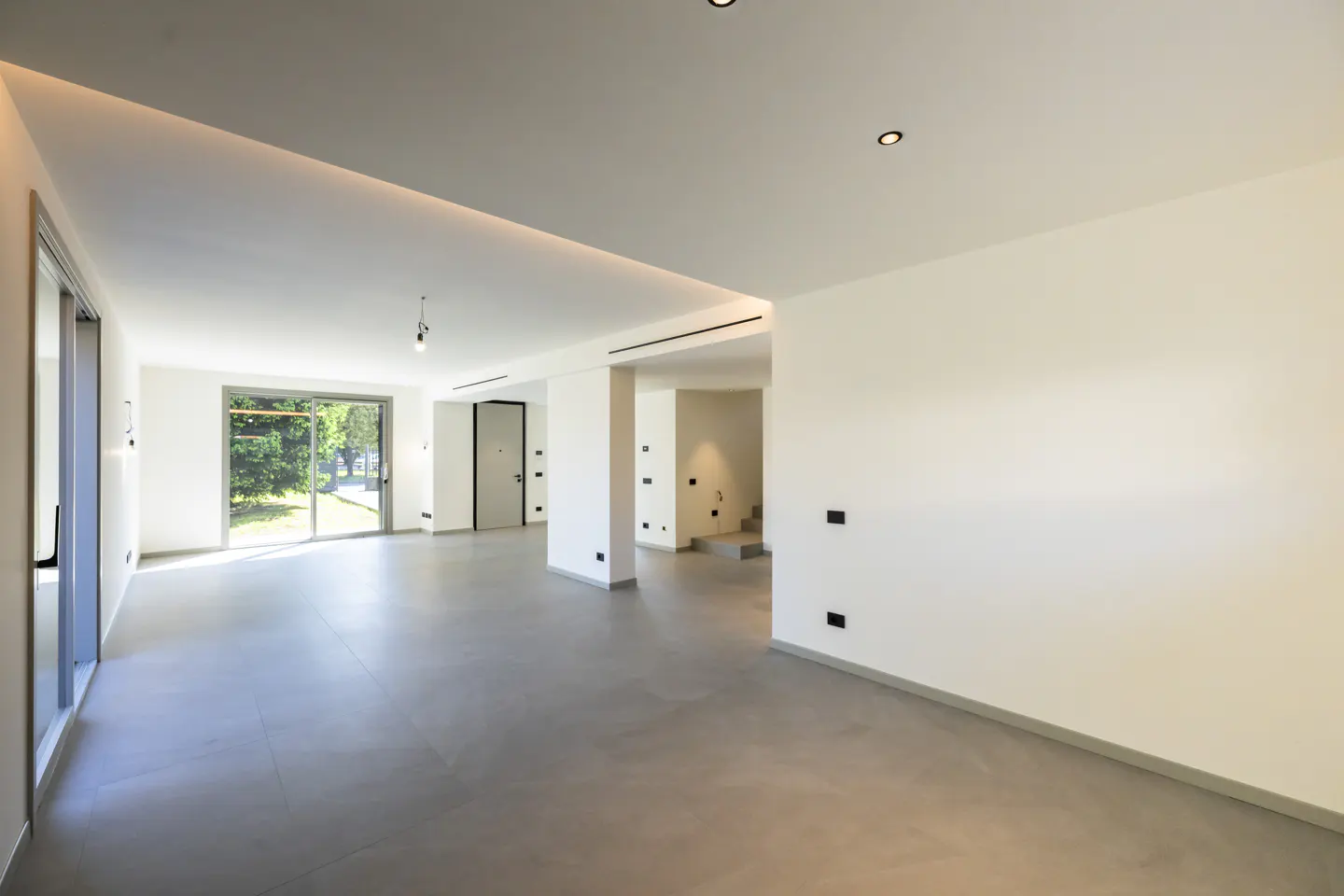 Bright, empty room with white walls, gray floors, and recessed lighting. A large window and door lead to the outside.