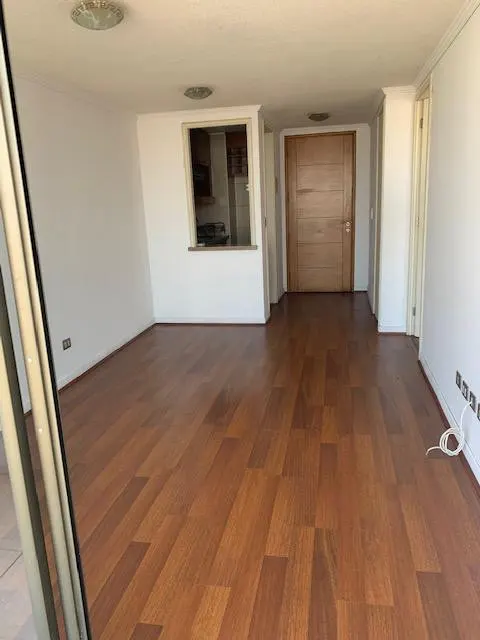 Empty room with hardwood floors, white walls, and a view into the kitchen through a window. A wooden door is visible.