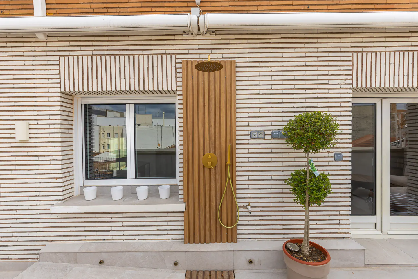Outdoor shower with wood paneling and gold fixtures against a brick wall. A small tree in a pot sits nearby.