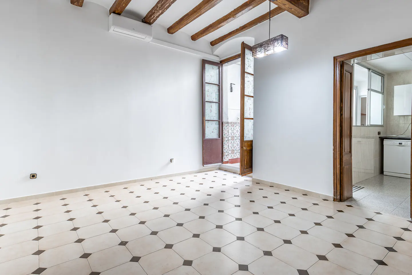 Empty room with white walls, tiled floor, and exposed wood beams. Open doors lead to a balcony and kitchen.