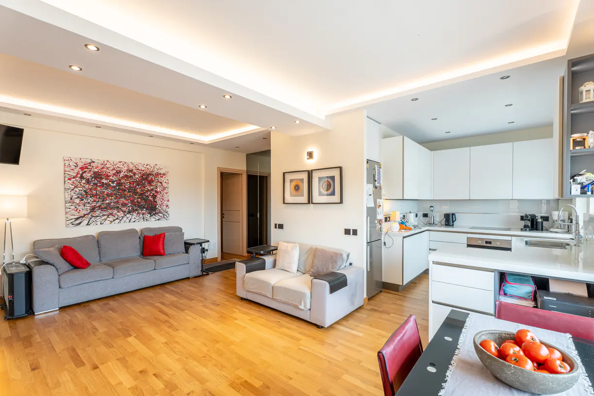 Open-plan living area with a grey sofa, white loveseat, modern kitchen, and hardwood floors, featuring a bowl of tomatoes.