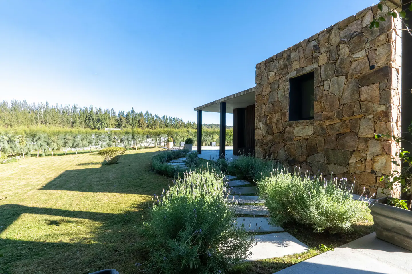 Stone-walled modern home with a stone path through lavender bushes, leading to a grassy lawn and a distant tree line.