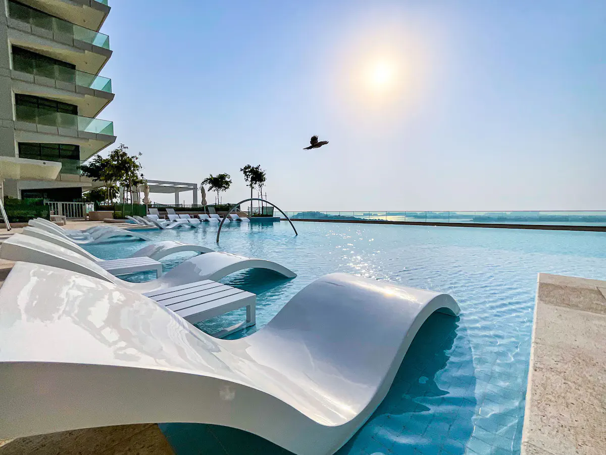 Infinity pool with white lounge chairs, overlooking the ocean under a sunny sky. A bird flies overhead. Condominium building on the left.
