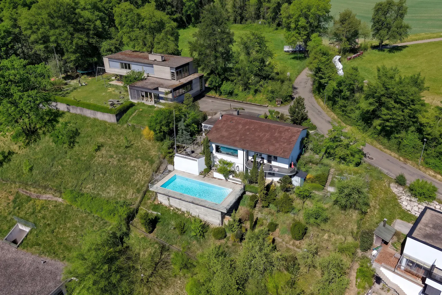 Aerial view of a white house with a brown roof and a blue swimming pool surrounded by green trees and grass.