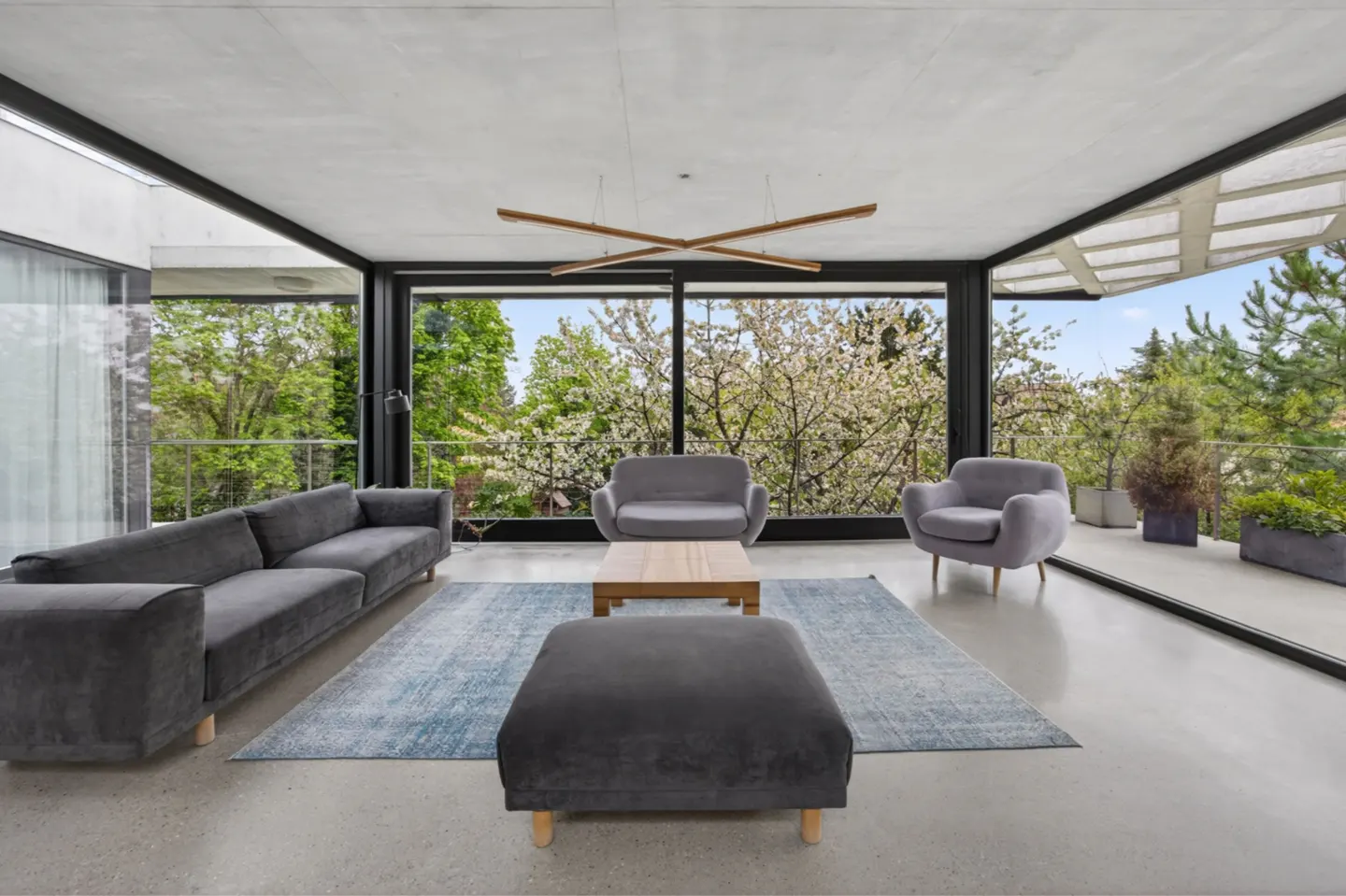Bright living room with gray sofa, chairs, and ottoman on a blue rug. Large windows offer a view of lush greenery.