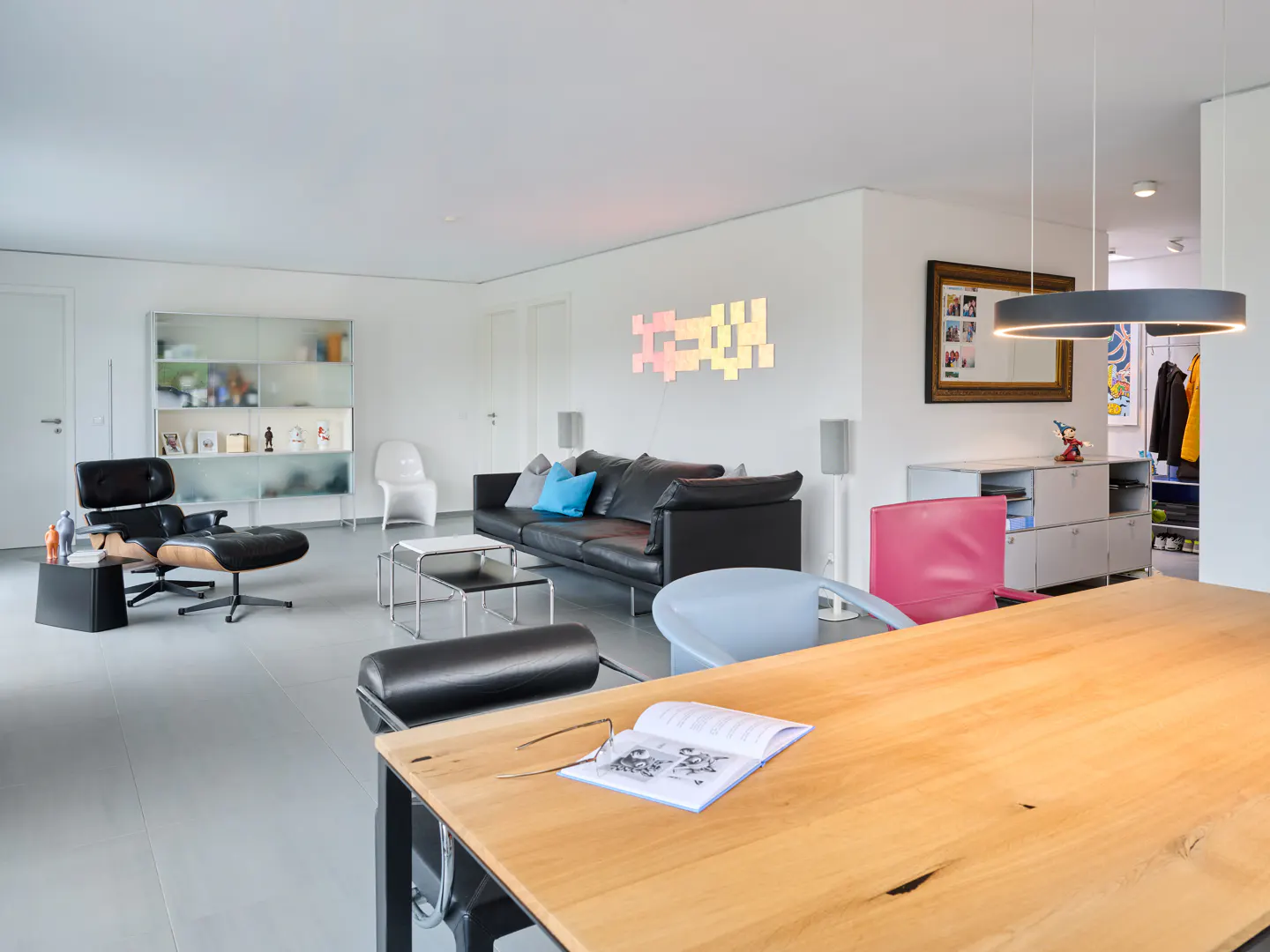 Bright, modern living room with a black leather sofa, Eames chair, and a large wooden table with a book and glasses.