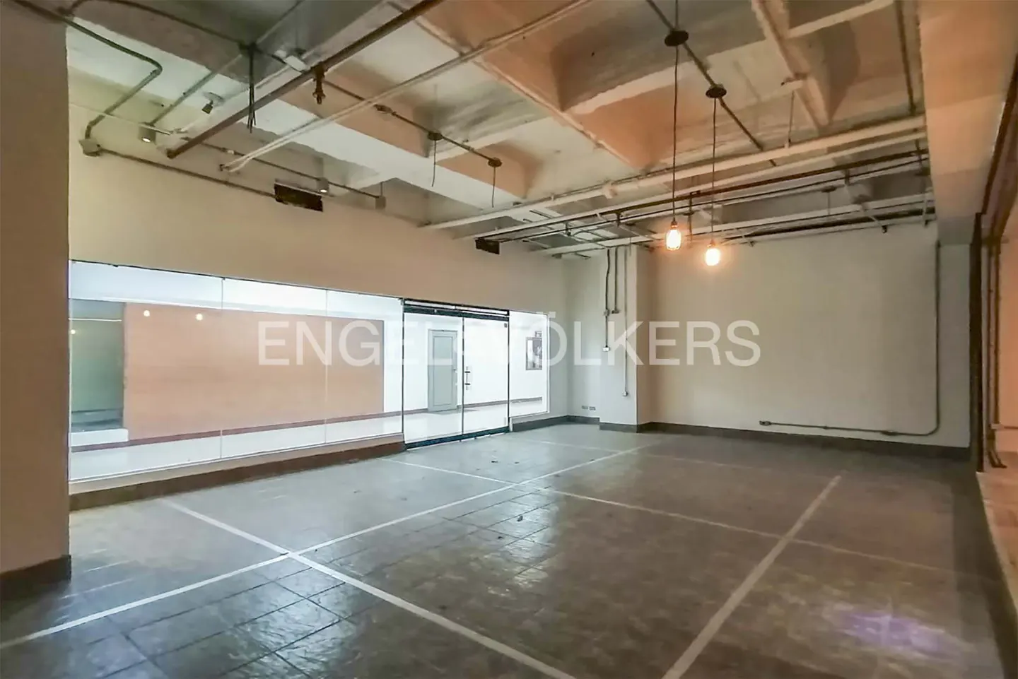 Empty commercial space with white walls, exposed ceiling pipes, and pendant lights. Floor is gray with white grid lines. Glass storefront on the left.