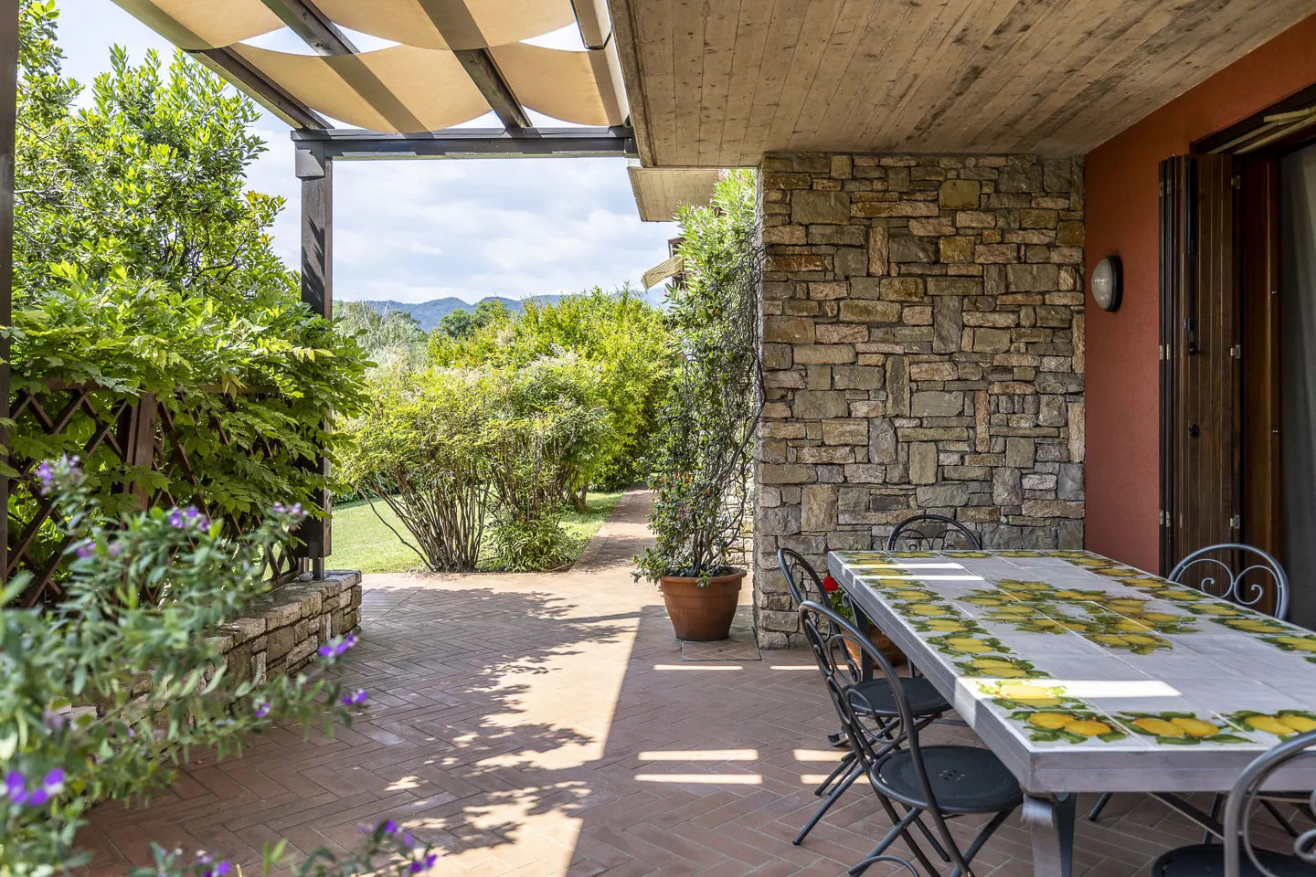 Outdoor patio with stone wall, terracotta floor, and a table with yellow lemon tile design. Green garden and trees in the background.
