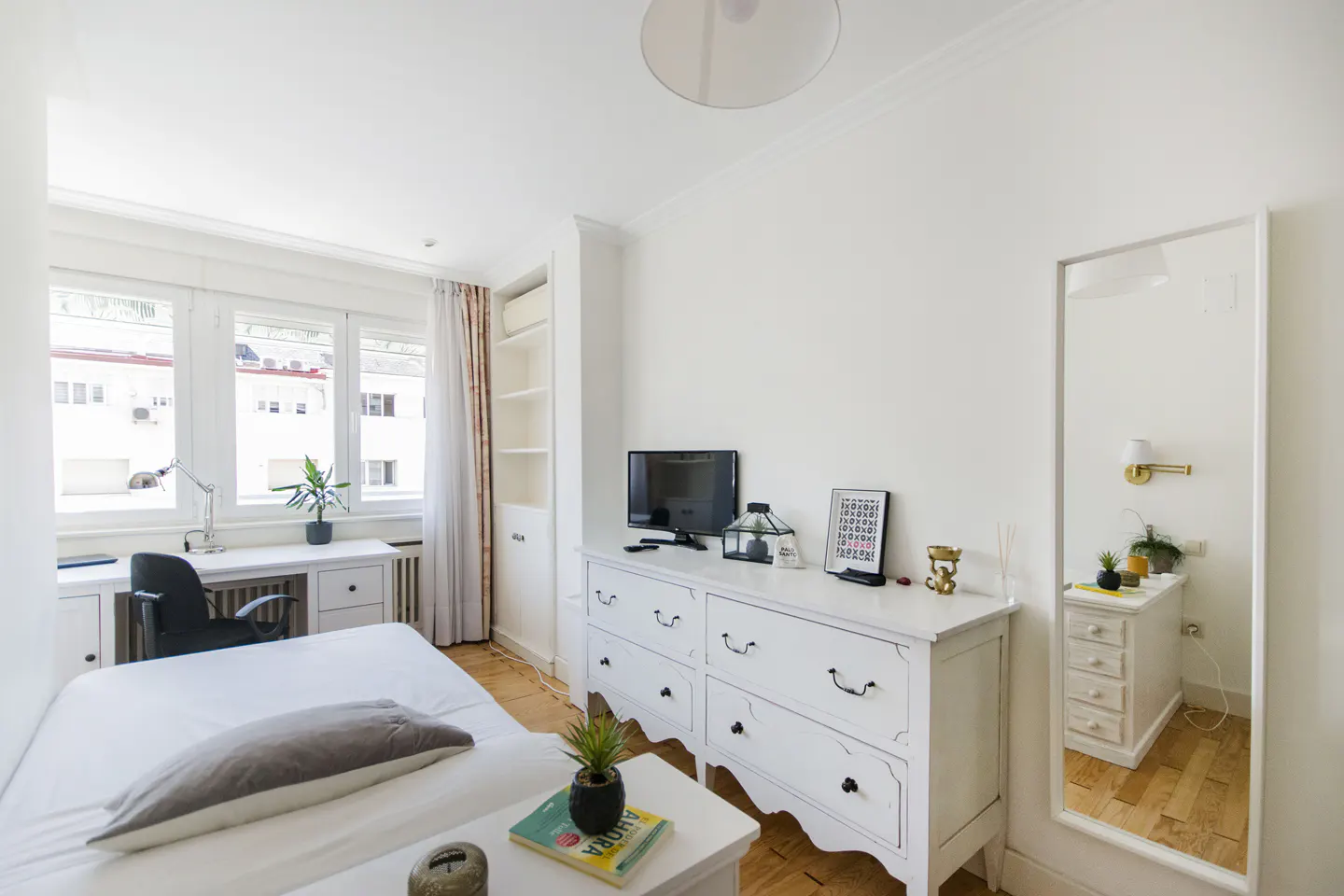 Bright bedroom with white walls, wood floors, and a large window. A bed, desk, dresser, and mirror furnish the space.
