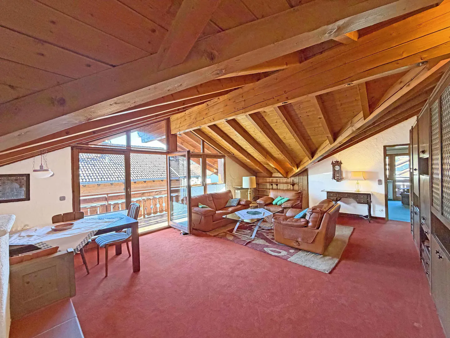 Attic living room with exposed wood beams, red carpet, leather sofas, and a dining table near a balcony with a view.