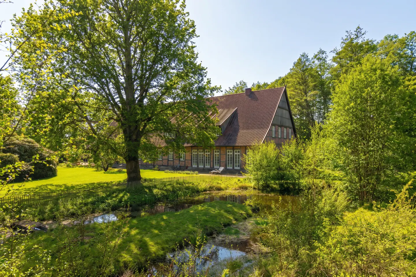 A large brick house with a tiled roof sits behind a lush green lawn, a large tree, and a small pond in a wooded setting.